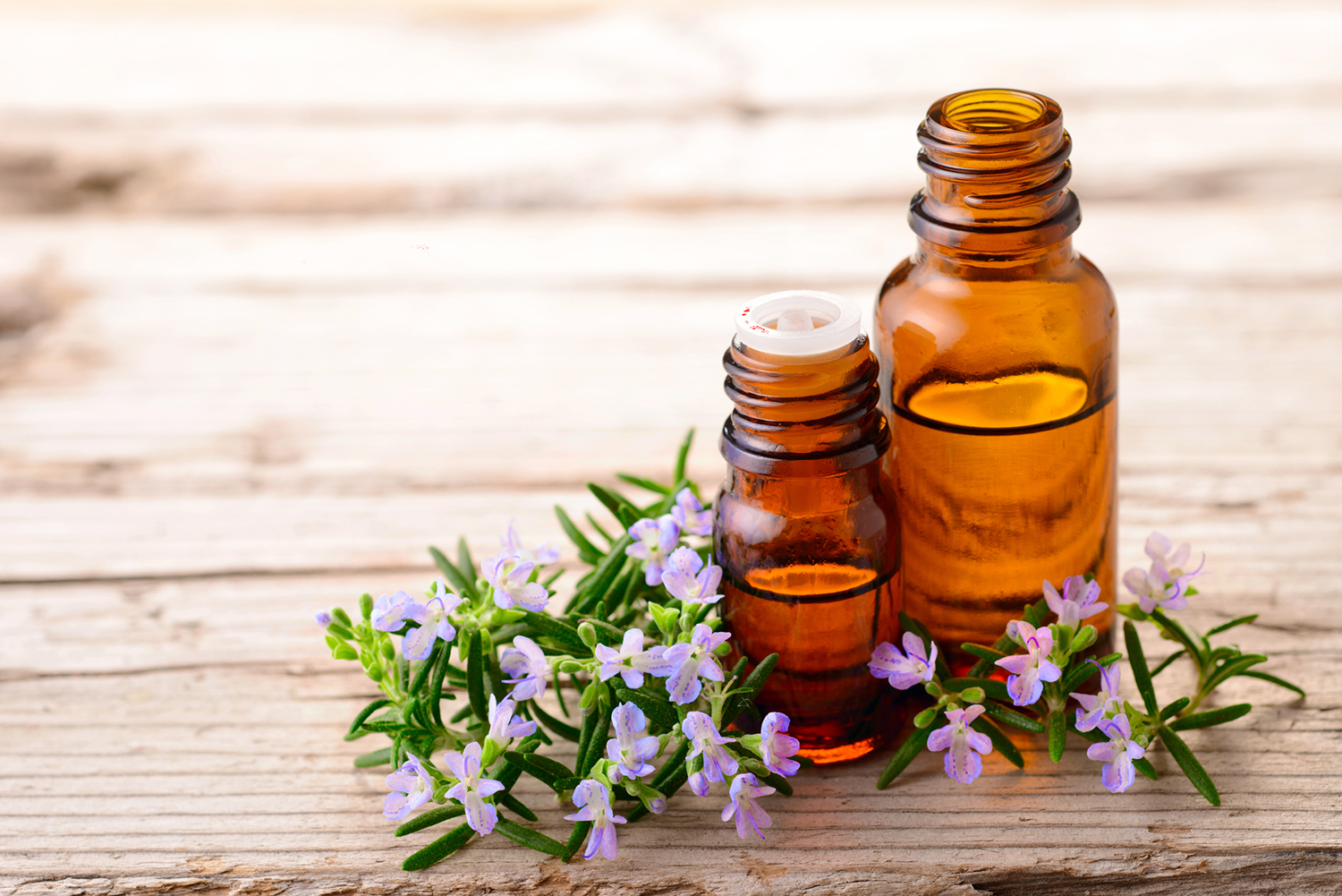 Blooming rosemary sprigs on a wooden table with essential oils in brown bottles.
