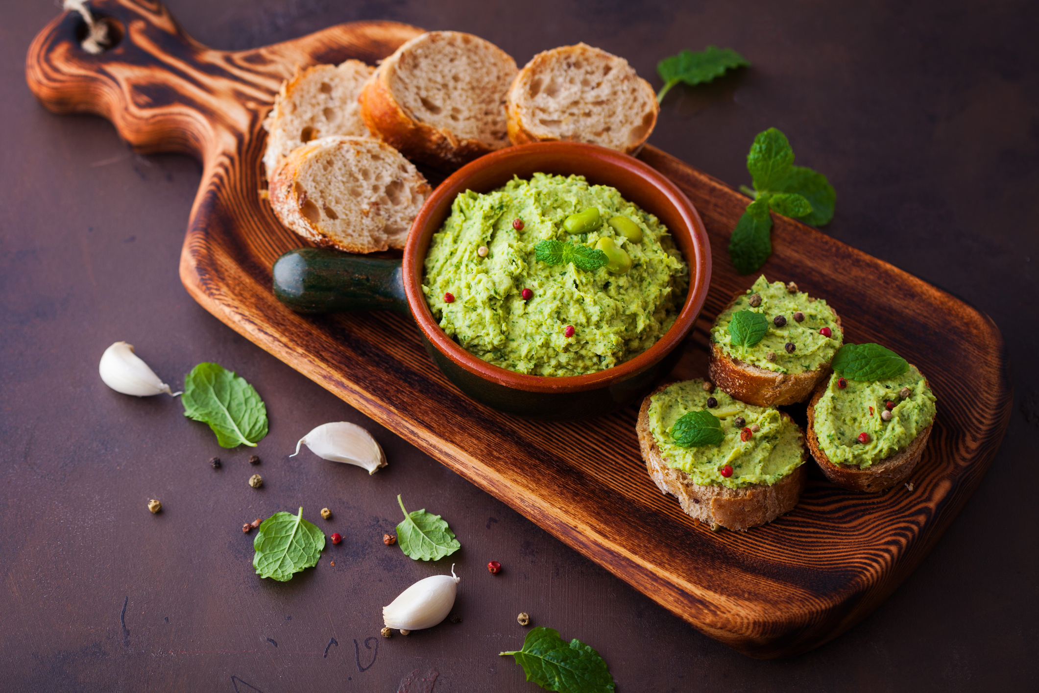 Bowl of fava hummus on a bread board along with bread rounds and three pieces of bread with fava hummus on them. 