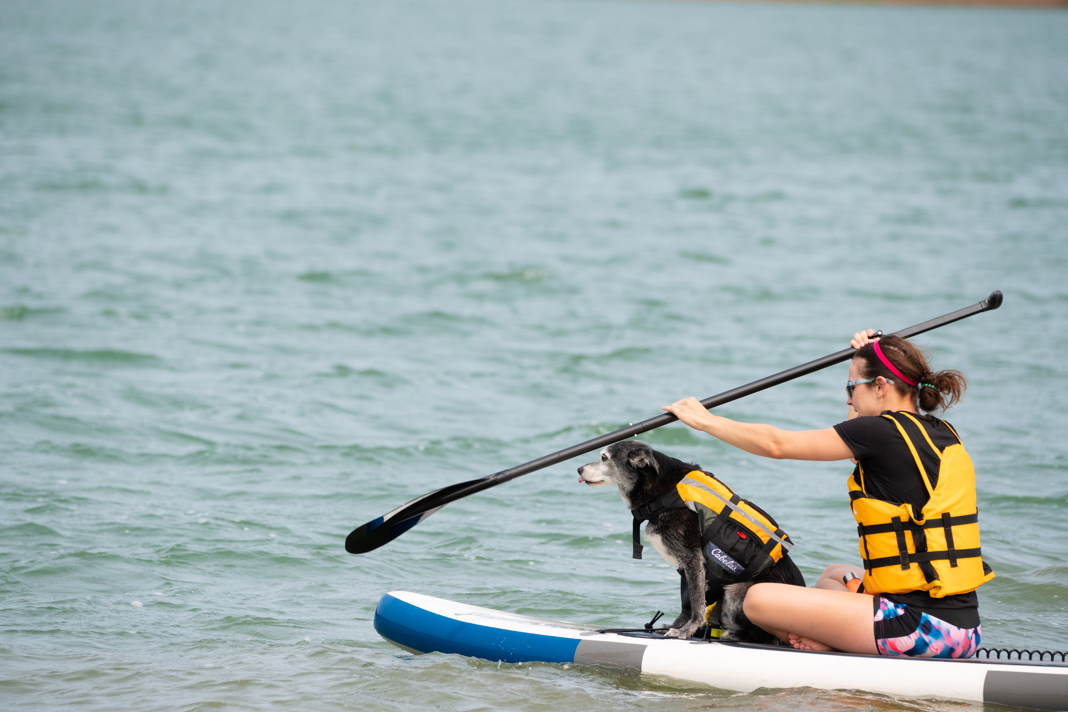 A woman with a yellow life vest is seated and paddling a paddleboat while an older black dog with a white muzzle, wearing a matching yellow safety vest, sits in front of her.