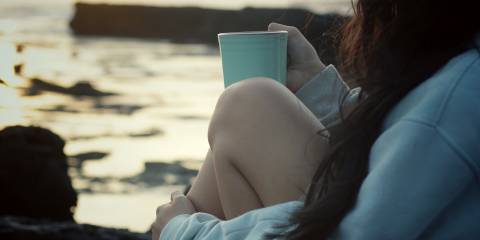 a young woman in comfy clothes drinking tea on a rocky shore