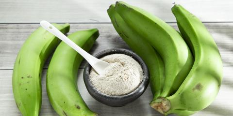 A small bowl of green banana powder with a spoon in it surrounded by green bananas,